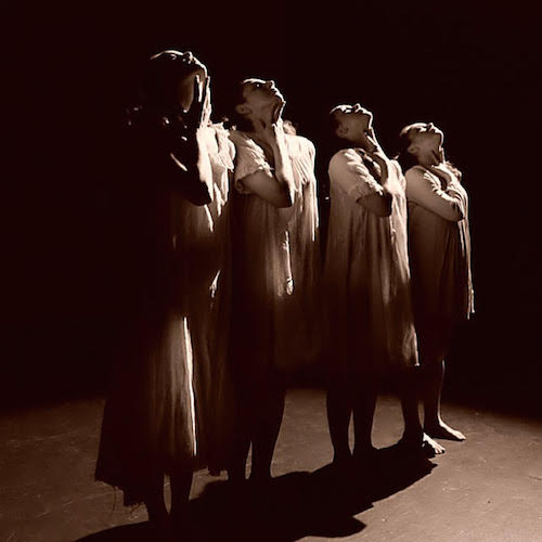 Four women stand in a straight line with their heads back and hands on their thoats.